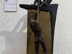 Eine Bronzeskulptur mit zwei Figuren, die eine sitzend, die andere hängend, steht auf einem Sockel mit einem Schild daneben.