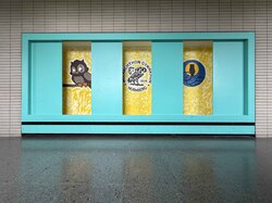 Drei Kunstwerke zum Thema Eule sind auf einem türkisfarbenen Hintergrund vor einer gefliesten Wand ausgestellt.