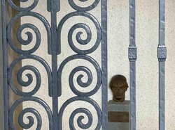 Ein dekoratives schmiedeeisernes Tor mit symmetrischen Wirbelmustern steht vor einer Mauer mit einer kleinen Statue.
