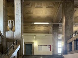 Ein architektonisches Interieur mit einem geometrischen Deckenmuster, einer großen Urnenskulptur und einer Treppe.