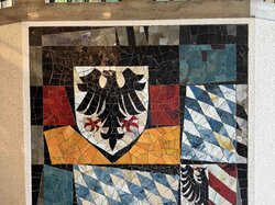 Ein Mosaik-Kunstwerk mit geometrischen Mustern und einem zentralen Emblem, das an einer Wand mit dekorativen Eisenfenstern angebracht ist.