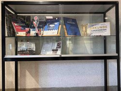Eine Glasvitrine enthält verschiedene Auszeichnungen, Urkunden und eine Trophäe.