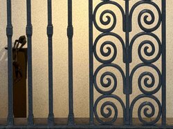 Ein schmiedeeisernes Tor mit symmetrischen Spiralmustern steht vor einer beigen Wand, durch deren Lücke die dunkle Silhouette einer Statue zu sehen ist.